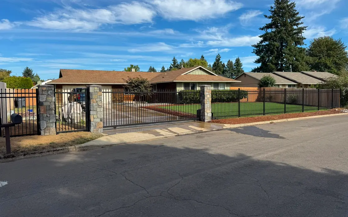 Wide street view of ranch home enclosed by black iron fence with stone columns. Fence & gate installation in Clackamas