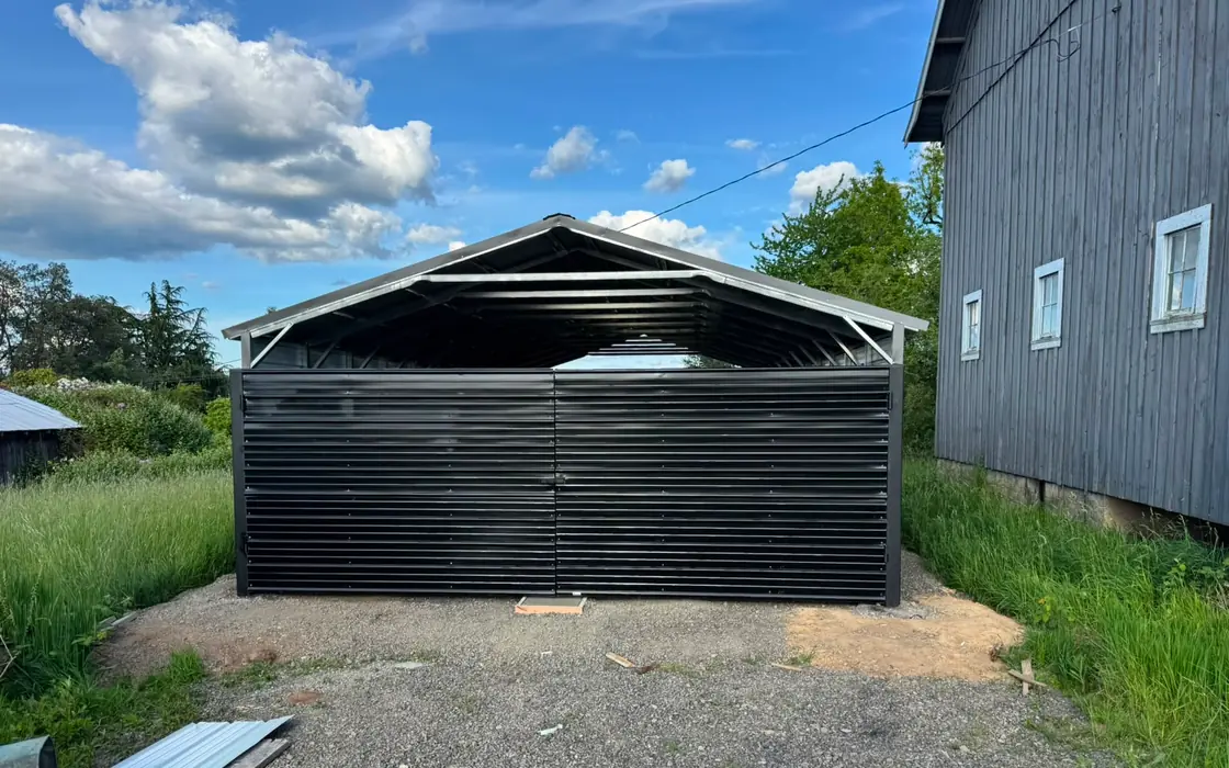 Black metal swing gate enclosing steel carport beside gray barn. Fence & gate installation in Fairview