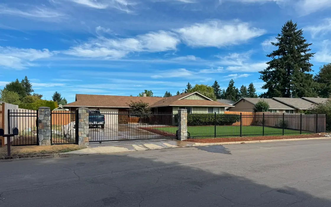 Wide view of ranch home with black iron fence, slide gate and stone columns on lawn. Fence & gate installation in Oregon City