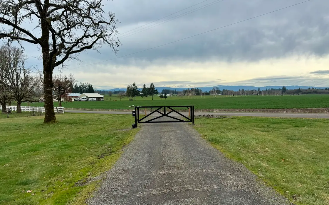 Wide view of closed black metal driveway gate across gravel drive with farm landscape. Fence & gate installation in Salmon Creek