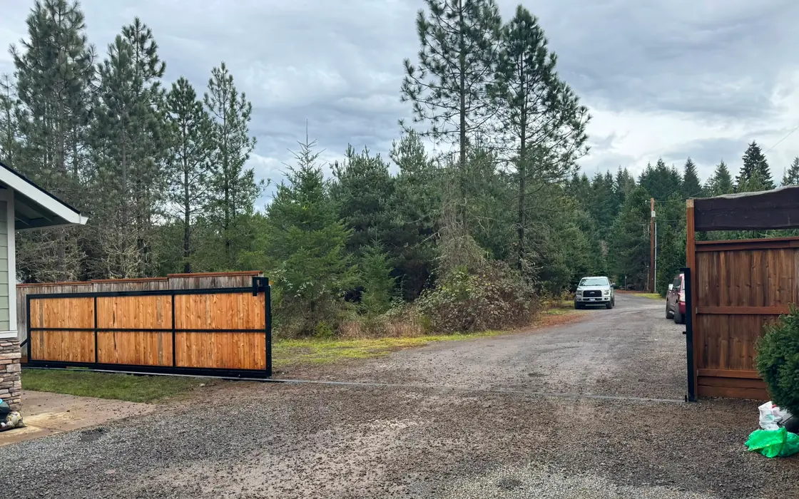 Rural driveway with open cedar rolling gate, gravel road and parked trucks. Fence & gate installation in Sandy