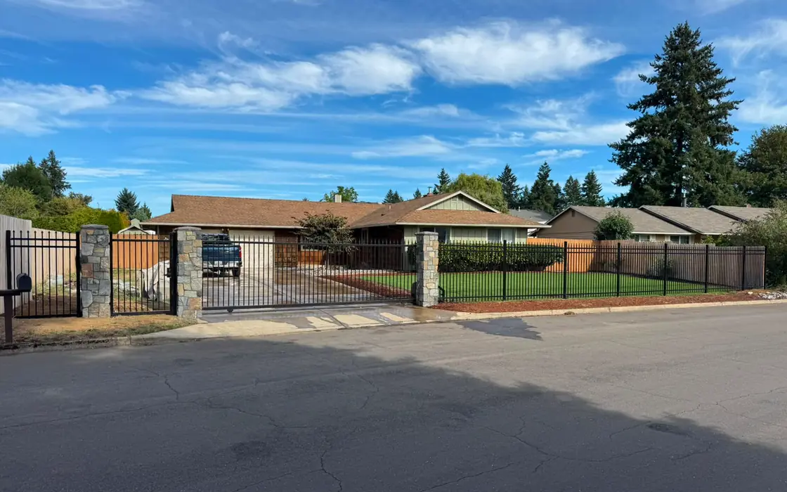 Street view of ranch house behind ornamental iron fence with stone columns and gate. Fence & gate installation in West Linn