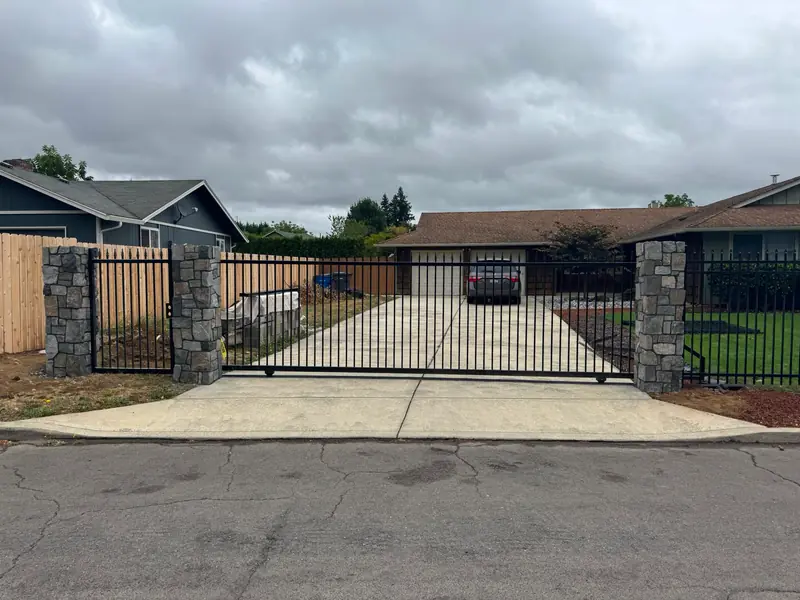 Automatic slide gate. Black iron slide gate closed across concrete driveway between stone columns, ranch home