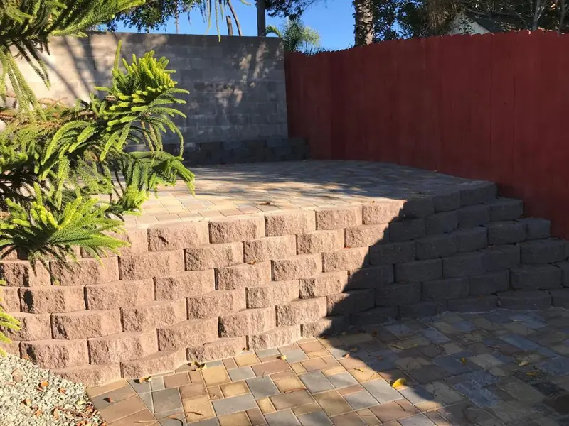 Stone-column entry. Finished paver patio with stacked-block retaining wall and red wood privacy fence