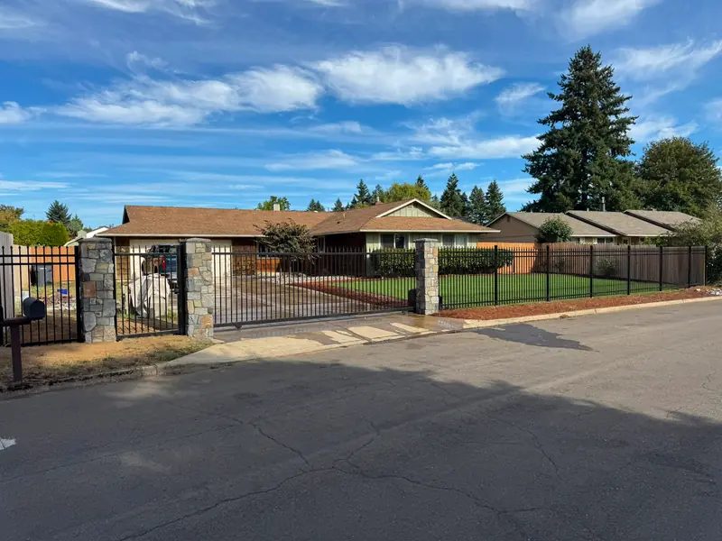 Wide street view of ranch home enclosed by black iron fence with stone columns